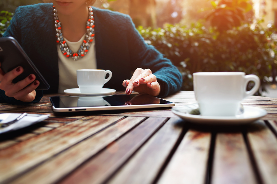 Cup of coffee on the foreground with elegant young woman using busy touch screen tablet at the coffee shop wooden table work break of business people flare sun light